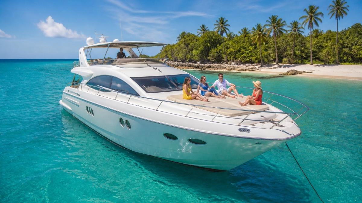 A white luxury yacht anchored in clear turquoise water near a tropical island, with a small group of guests relaxing on the sun deck under a blue sky